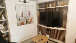 A modern living room with a flat-screen TV, two round tables, a black chair, a painting, and a decorative wall panel.