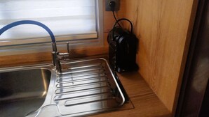 A compact kitchen with a stainless steel sink, a window with blinds, and a black kettle on the counter.
