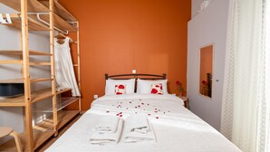 Ein ordentlich bezogenes Bett mit weißen Leinen und roten Rosenblättern, ein Holzregal mit einem weißen Handtuch und ein Spiegel an der Wand.