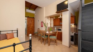 Quarto compacto com cama, mesa de jantar e cadeiras, cozinha com armários e eletrodomésticos, e uma televisão instalada acima da mesa.