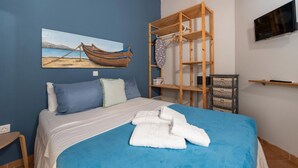 Une chambre à coucher avec un lit, un tableau représentant un bateau accroché au mur, une étagère en bois et un téléviseur fixé au mur.