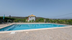 Une piscine entourée d’une terrasse en pierre, un petit bâtiment avec un toit jaune et un paysage montagneux en arrière-plan.