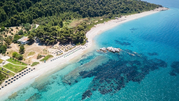 Vue aérienne d’une zone côtière avec une plage, des eaux d’un bleu limpide et une colline boisée.