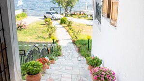 Un sendero de piedra conduce a un edificio blanco con contraventanas de madera, plantas en macetas y vistas al mar.