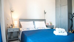 Una cama bien hecha con una colcha azul y almohadas blancas, una lámpara de noche, una mesita de noche con un jarrón y una lámpara, y un armario gris.
