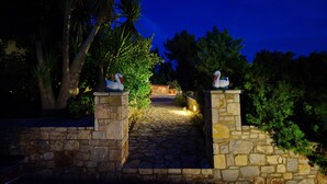 A stone pathway leading to a building entrance, flanked by stone pillars with swan statues, surrounded by greenery. - Kamelia Villa 1 in Alonissos (Votsi)