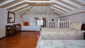 A bedroom with two beds, wooden flooring, a sloped ceiling with exposed beams, a desk with a chair, and a window with a view of greenery. - Kamelia Villa 1 in Alonissos (Votsi)