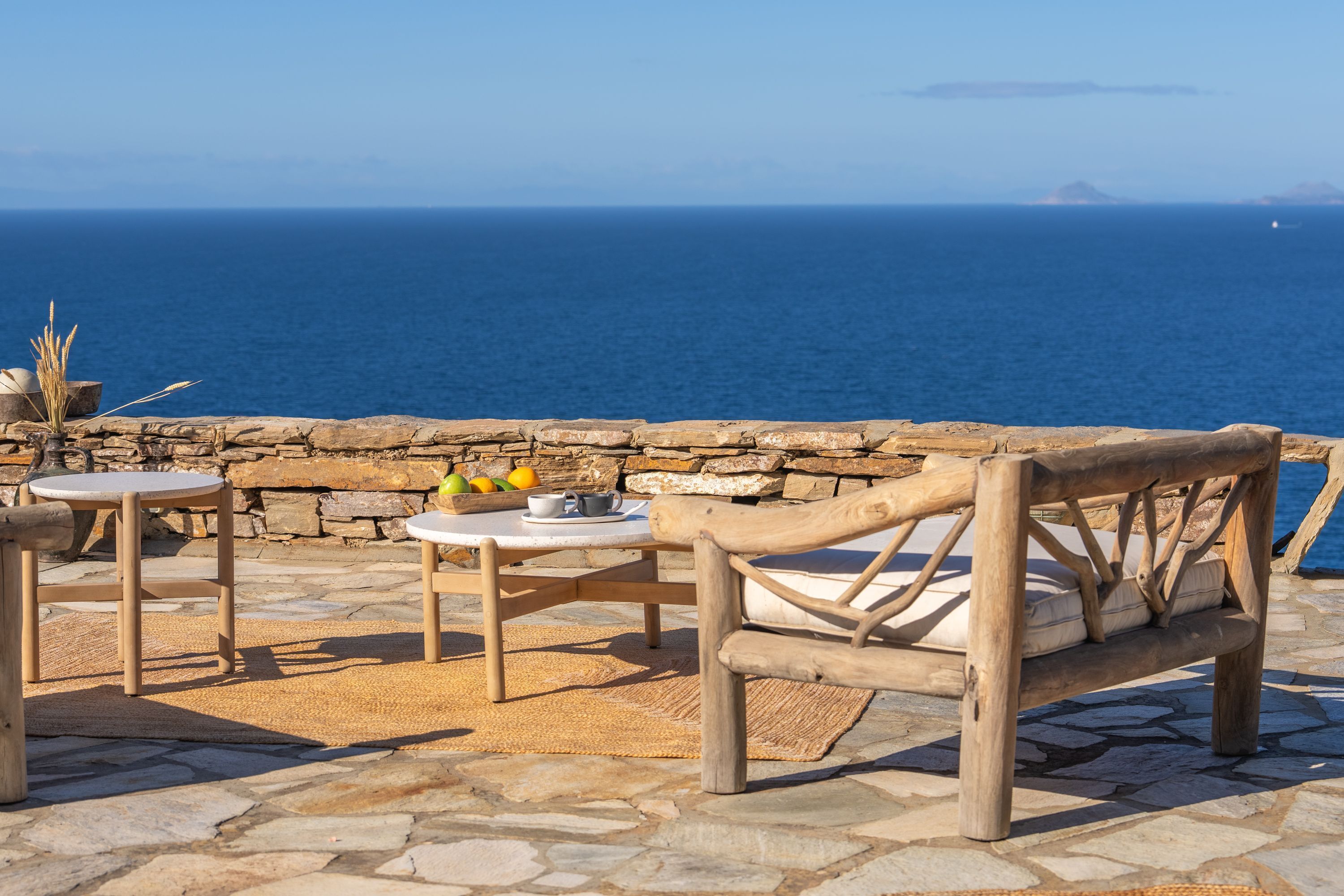 Un patio de piedra con un banco de madera y dos mesas redondas, una con un frutero y una tetera.