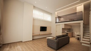 A modern living room with a sofa, a flat-screen TV, and a kitchen area visible through an open door.