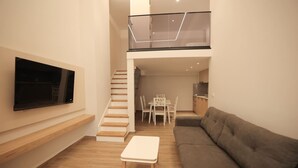 A modern living room with a flat-screen TV mounted on the wall, a staircase leading to an upper level, a dining area with a table and chairs, and a sofa. - Duplex City 05 in Durrës (Durrës)