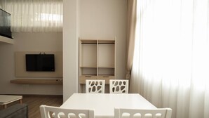 Uma sala de estar moderna com uma mesa e cadeiras brancas, uma TV de tela plana na parede e uma estante embutida.
