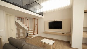 Sala de estar moderna com um sofá cinza, uma mesinha branca, uma TV na parede e uma escada que leva a um andar superior.