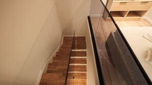 Un escalier moderne avec des marches en bois et une rampe en verre, menant à une chambre à coucher équipée d’un lit et de rangements intégrés.