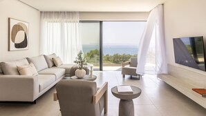 A modern living room with a sofa, armchair, and coffee table, a flat-screen TV, and a large window with a view of the sea. - Syvana Exquisite Villas Complex IV (Syvota)