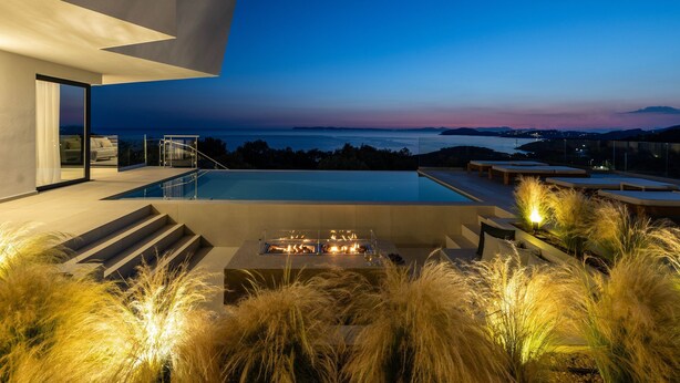 A modern outdoor pool area with a view of the sea at dusk, featuring a fire feature and outdoor seating. - Syvana Exquisite Villas Complex IV (Syvota)