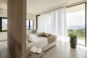 A bedroom with a bed, a chair, a potted plant, and a view of the sea. - Syvana Exquisite Villas Complex IV (Syvota)
