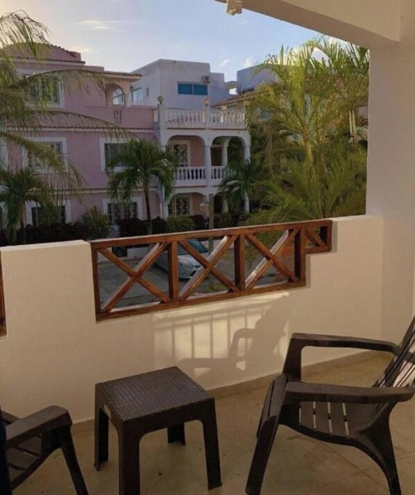 A balcony with a wooden railing, two chairs, and a small table. In the background, there are buildings with balconies and palm trees. - apartment for rent (Bayahíbe)