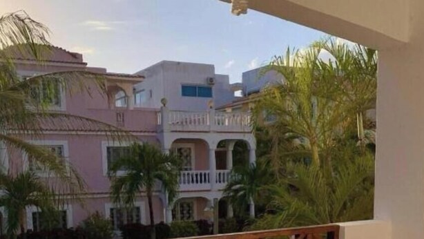 Un balcón con barandilla de madera, dos sillas y una mesita. En el fondo, edificios con balcones y palmeras.