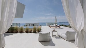 A modern outdoor lounge area with white cushions, a small table, and a view of the sea and mountains.