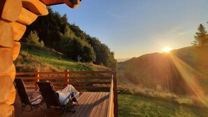 Een houten terras met twee loungestoelen en een persoon zittend, met uitzicht op een grasheuvel in de zonsondergang.