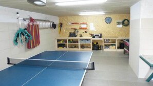 Een kamer met een tafeltennistafel, planken vol diverse spullen, een dartbord en een plafondventilator.