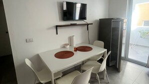 Ett modernt kök med ett vitt bord, två vita stolar, en svart tv som hänger på en vägghylla och ett kylskåp.