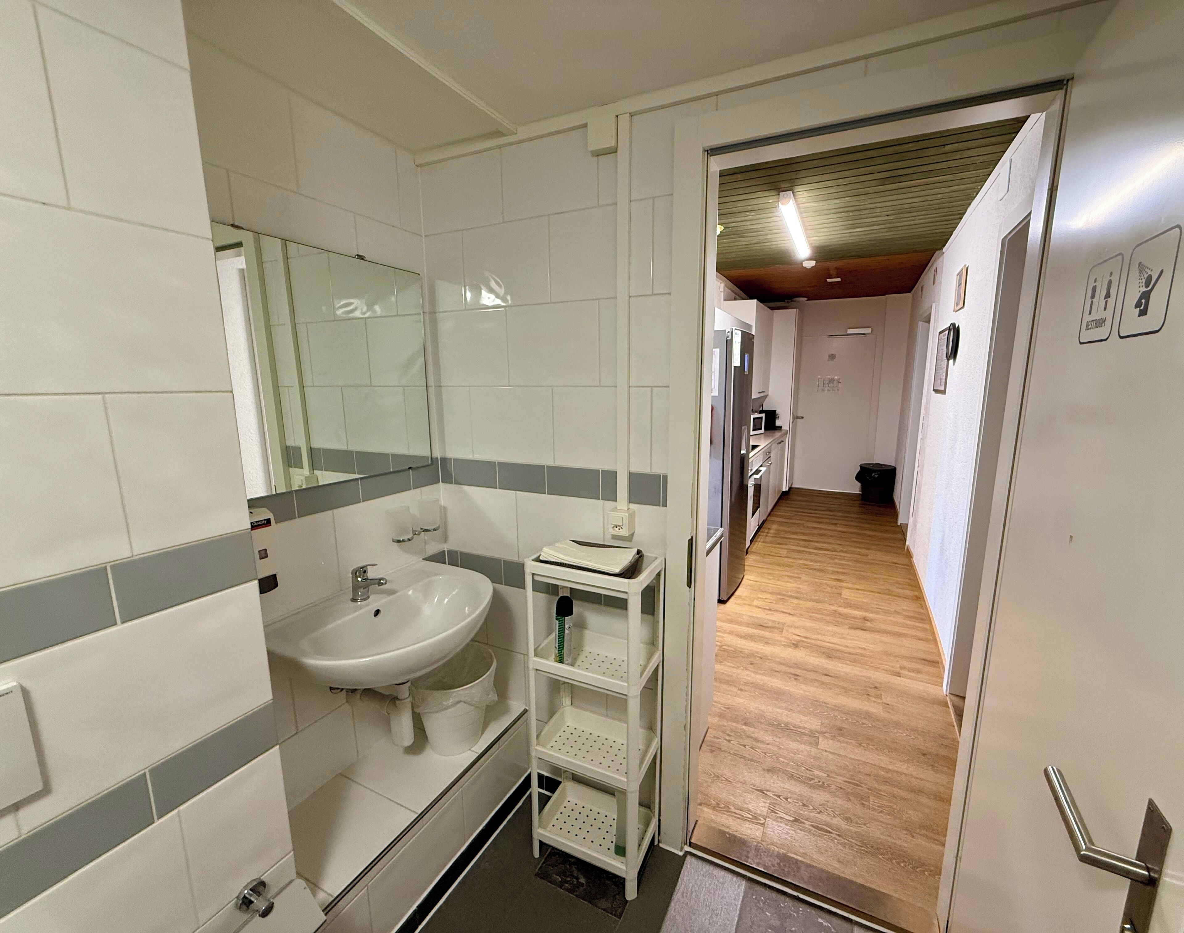 A narrow hallway with a bathroom on the left, featuring a sink, mirror, and tiled walls. A wooden floor leads to a kitchen area with appliances and a microwave.