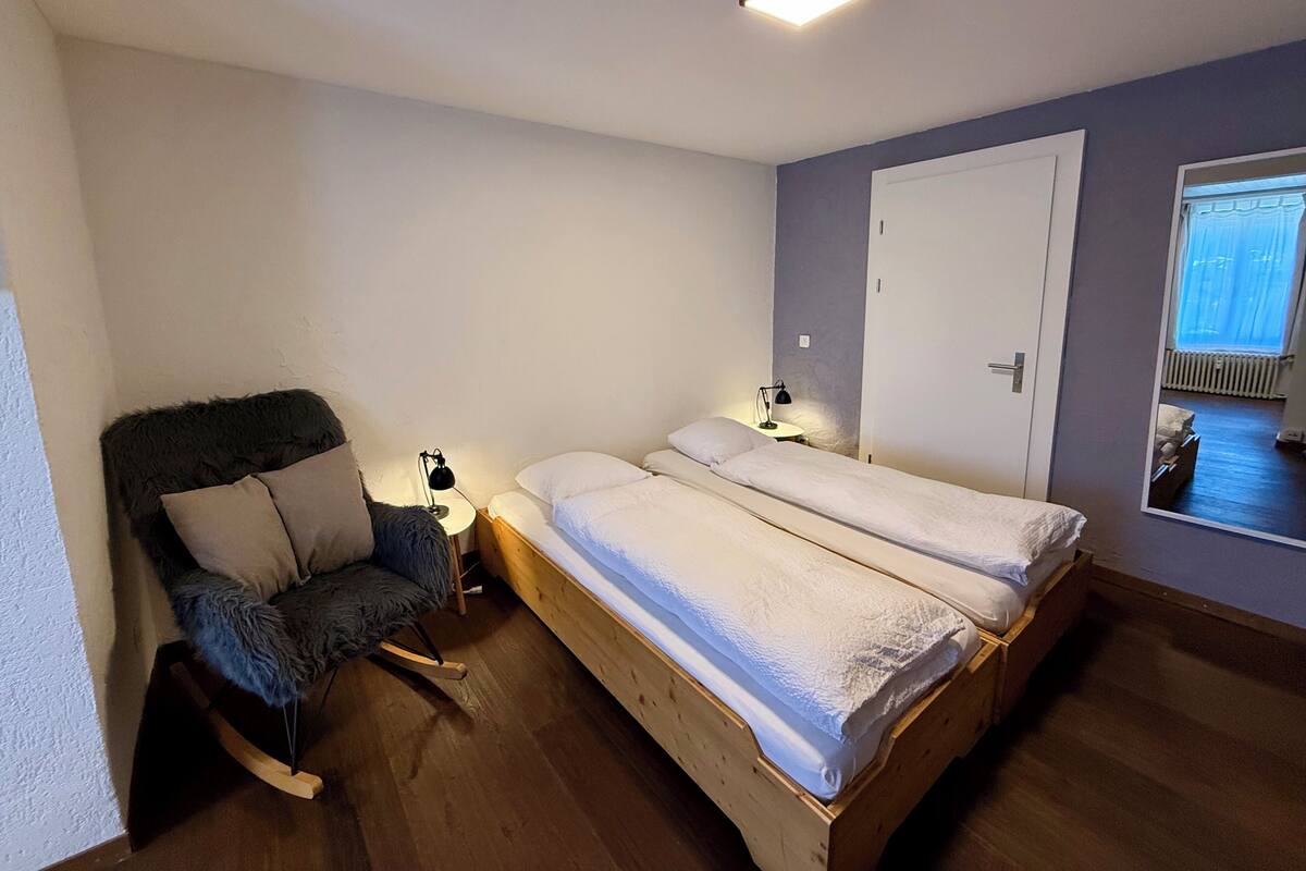 Una habitación pequeña y bien iluminada con dos camas individuales, una mecedora, una mesita de noche con lámpara y una puerta con espejo.