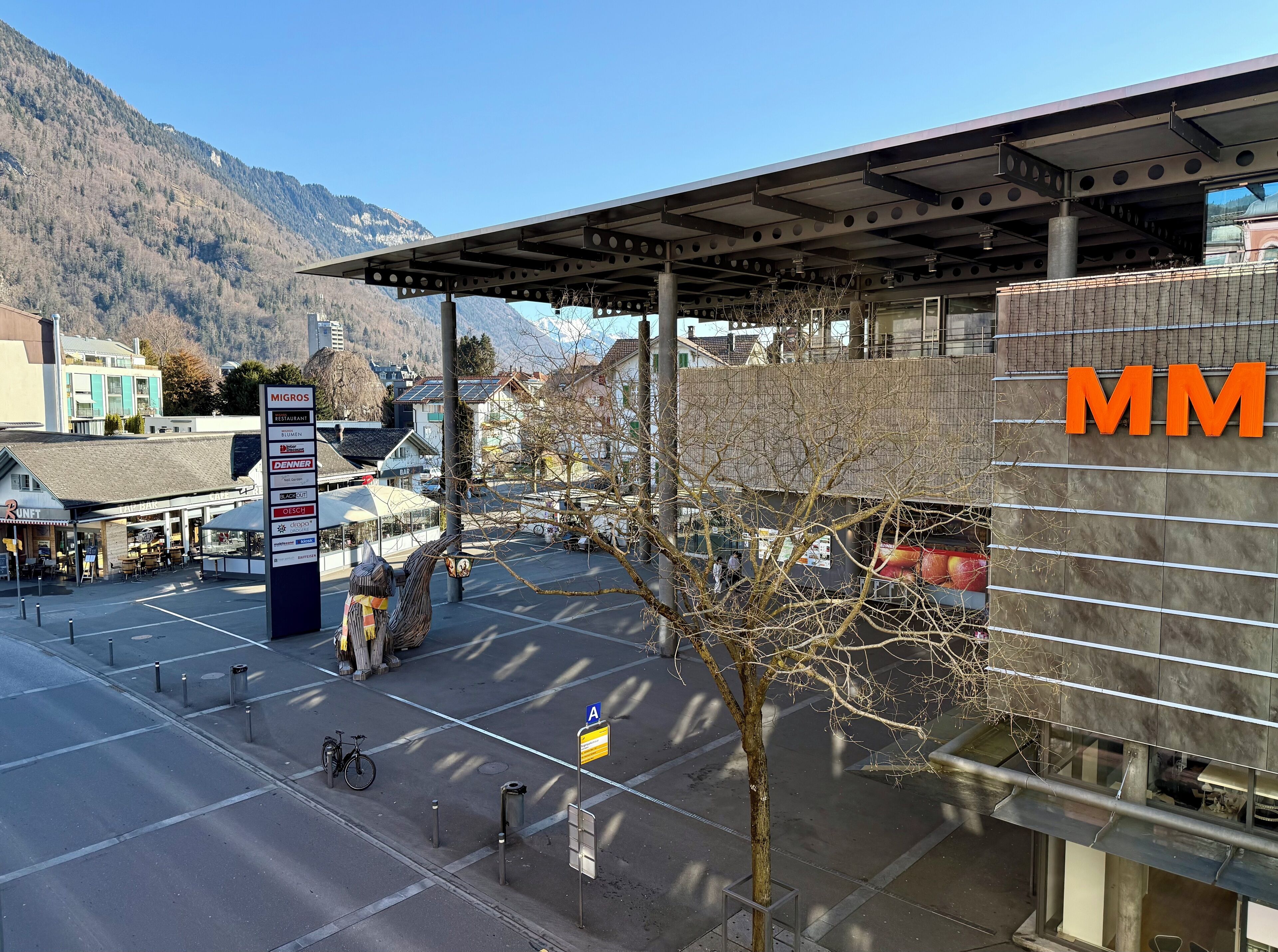 A modern building with a large 'MM' sign, a covered outdoor area, and a mountain backdrop.
