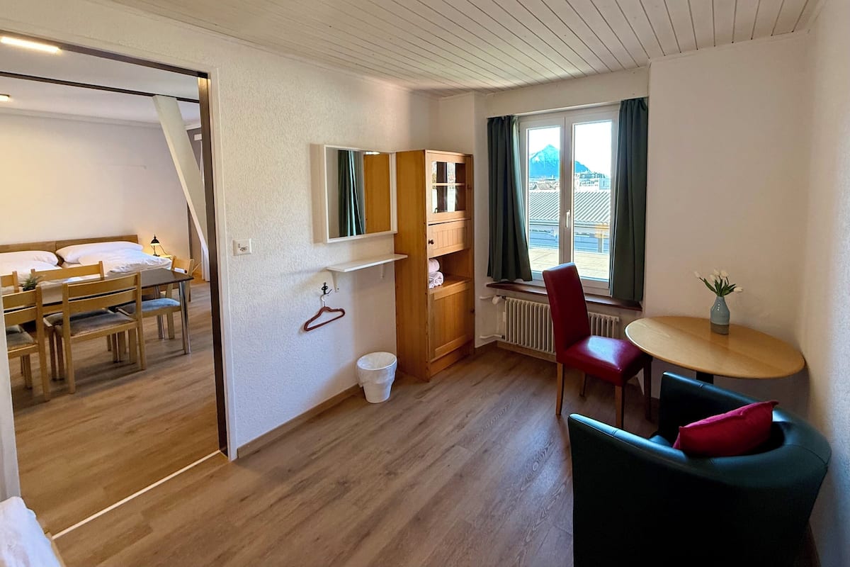Habitación de hotel con suelo de madera, zona de comedor con mesa y sillas, cama, espejo, pequeño armario, ventana con cortinas y una silla roja.
