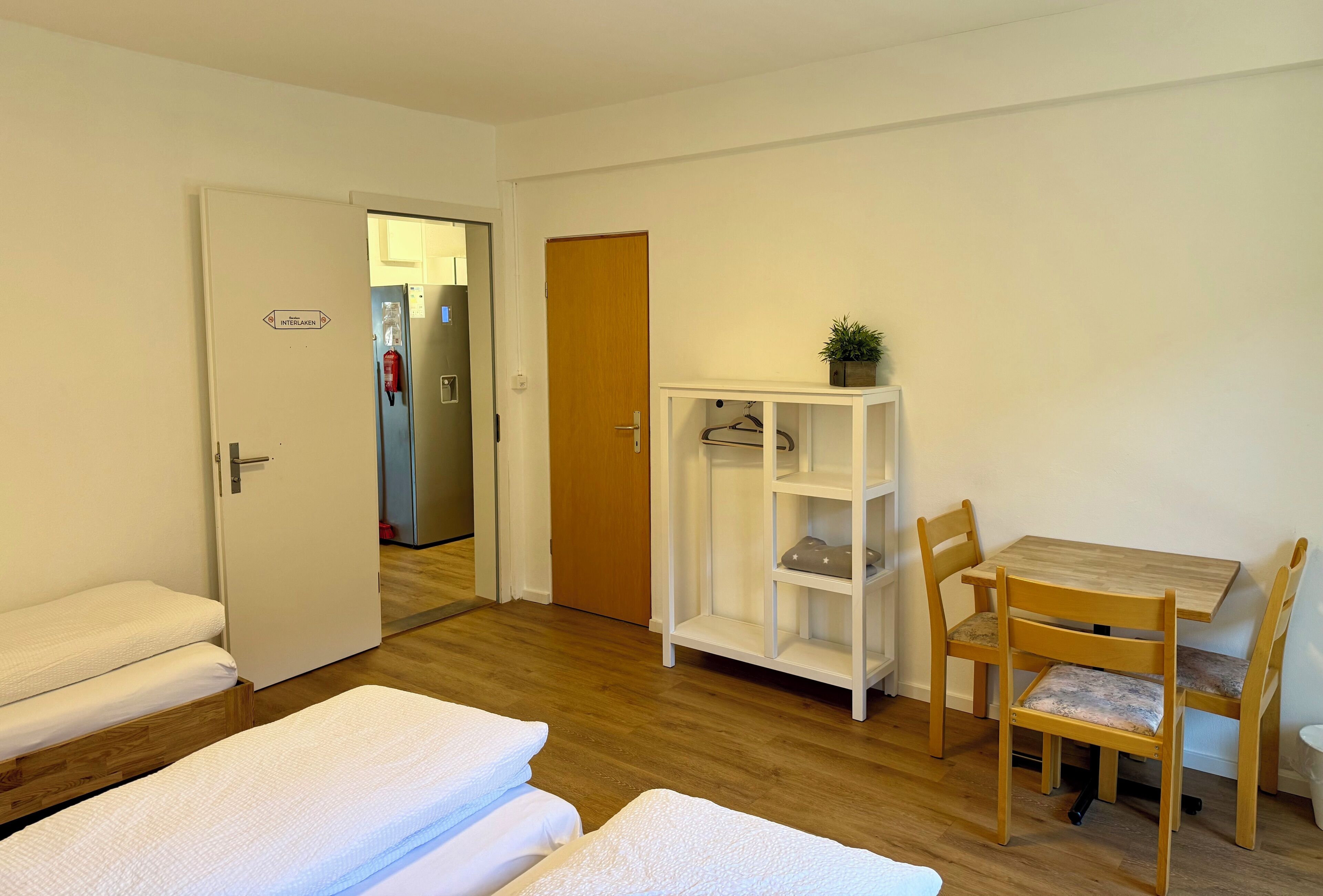 Habitación de hotel con una cama, una pequeña mesa con sillas, un armario blanco y una puerta que da a otra habitación.