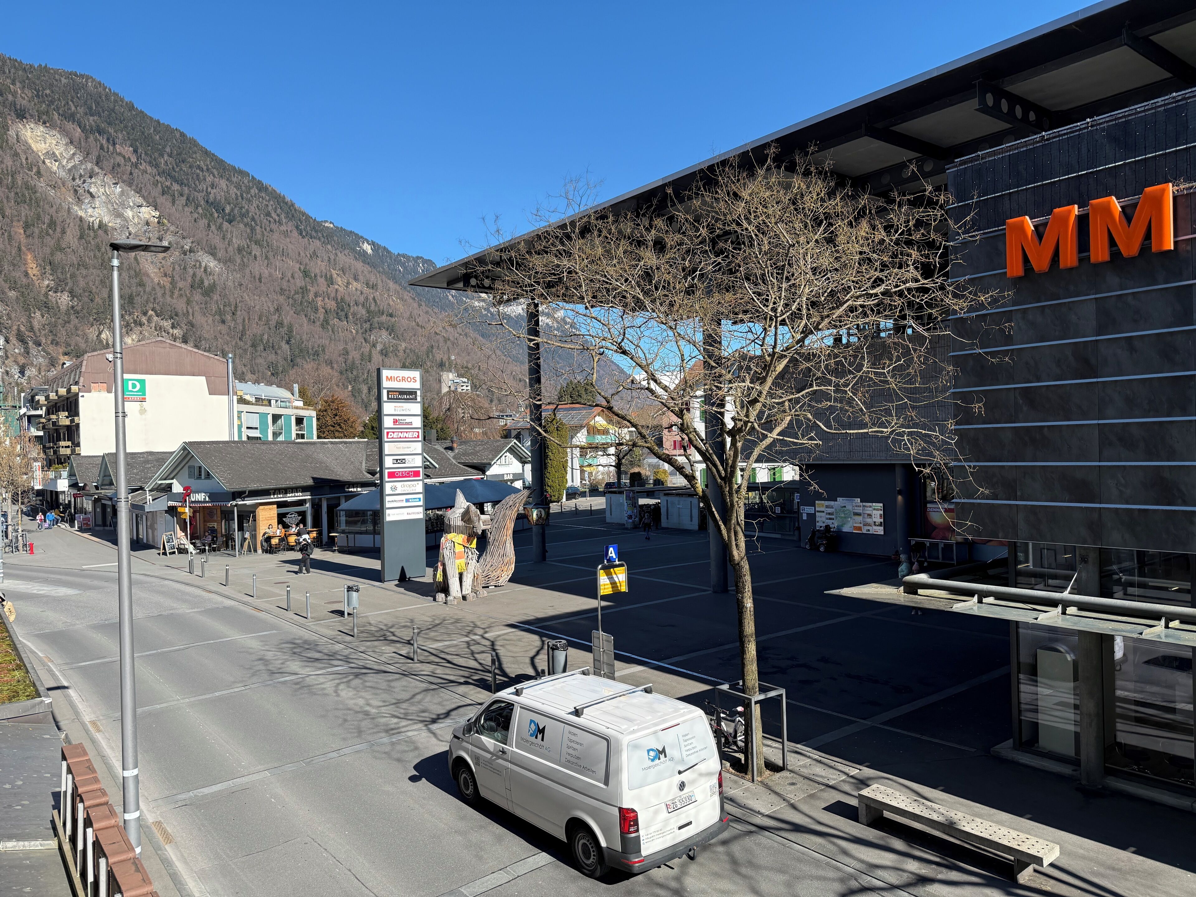 A commercial area with a white van parked in front of a building with 'MM' signage, a signpost with business names, and a mountainous backdrop.