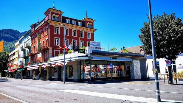 A street view with a red building featuring a 'Café' sign and a storefront with a 'P' parking sign. - DownTown Apartments (Interlaken)
