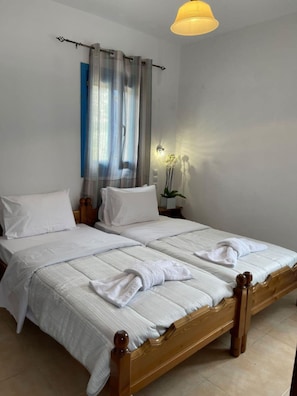 Un lit double avec du linge de lit blanc et une tête de lit en bois, une table de chevet avec une lampe et une fenêtre avec des rideaux légers.