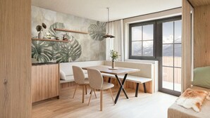 A modern living room with a wooden floor, a dining table with chairs, a bench, a large window with a view of mountains, and a wall with a tropical leaf mural. - Apartment mit 1 Schlafzimmer (Kaprun)