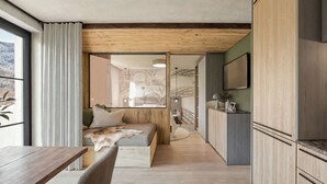 A modern interior with a wooden ceiling, a seating area with a fur throw, a kitchen with a TV, and a large window with a view of trees. - Apartment mit 1 Schlafzimmer (Kaprun)
