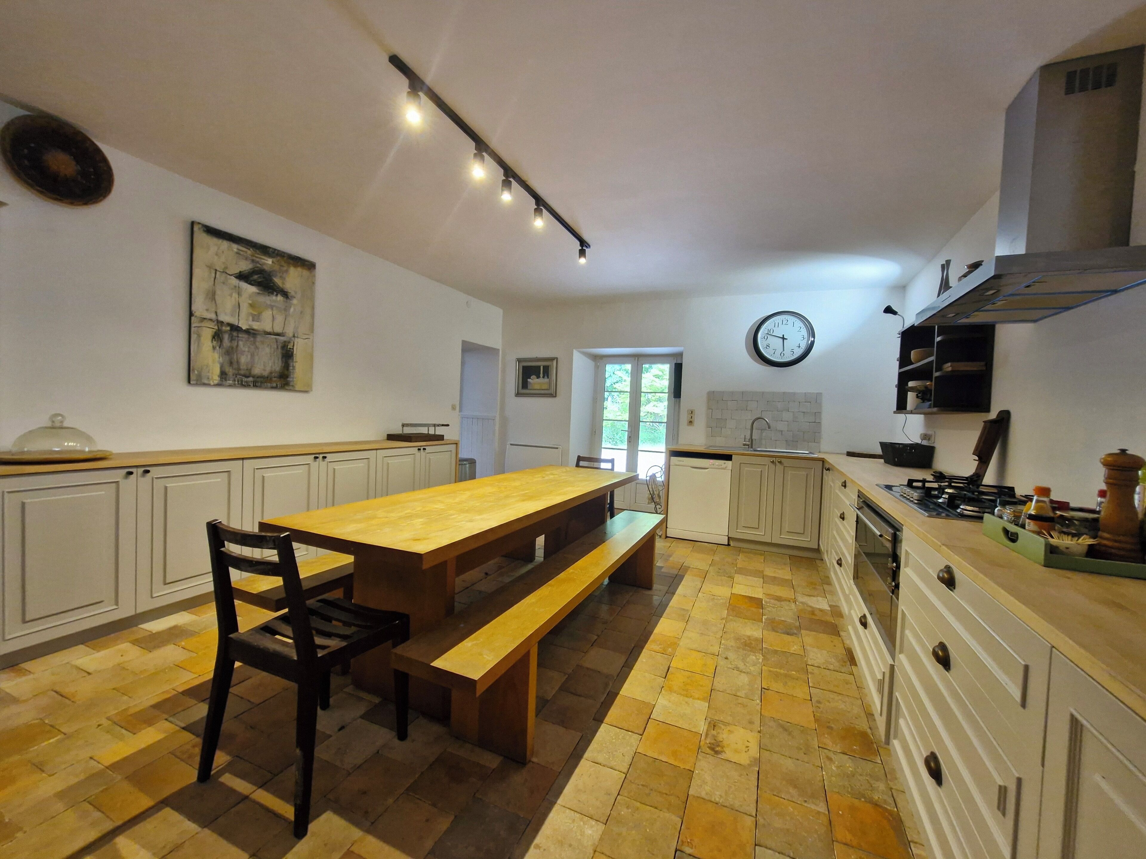Een keuken met een houten tafel en bank, witte kastjes, een klok en een raam.