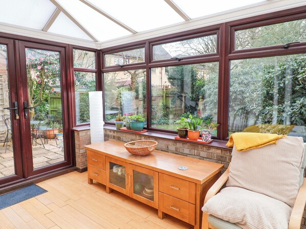 A conservatory with a wooden bench, a wicker chair, and a view of a garden through large windows.