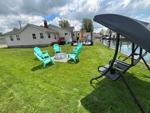 A backyard with a lawn, a metal fire pit, and four turquoise Adirondack chairs arranged around it. - Cozy 2 bedroom corner canal cottage in gorgeous Algonac with AC. (Algonac)