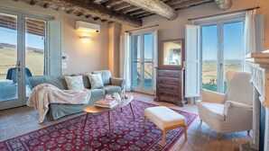 A cozy living room with a sofa, a coffee table, and a chair. There are large windows with a view of the countryside.