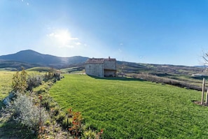 Un paysage rural avec une maison en pierre sur un champ verdoyant, entouré de collines et sous un ciel dégagé.