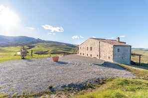Un bâtiment en pierre avec une allée de gravier, entouré de collines verdoyantes et sous un ciel dégagé.