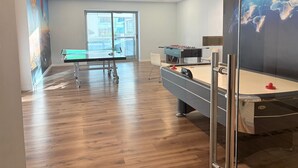 Una habitación con mesa de ping pong, mesa de futbolín y mesa de billar. Un ventanal grande proporciona luz natural.