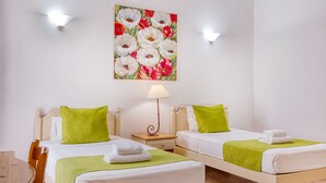Ein Hotelzimmer mit zwei Betten, jedes mit einem grünen Kopfkissen und einem weißen Handtuch. An der Wand hängt ein Blumenbild, auf einem hölzernen Nachttisch steht eine Lampe und es gibt einen Holzstuhl.