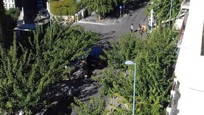 Vista de uma rua arborizada, com carros estacionados e edifícios ao fundo.