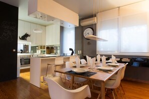 A modern kitchen with white cabinets, a dining area with a wooden table and white chairs, and a large window with a clock on the wall. - ACROPOLITANA 2 (near Acropolis and museum) (Athina)