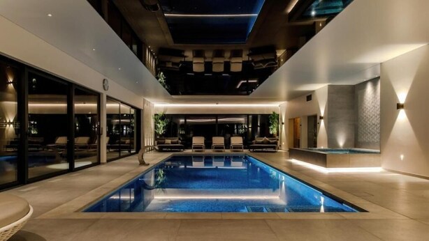 Un área de piscina interior moderna con una piscina azul cristalina, sillas de descanso e iluminación empotrada en el techo.