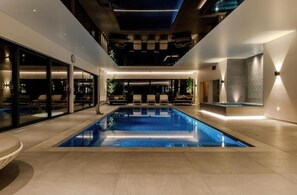 Ein moderner Poolbereich im Innenbereich mit einem klaren, blauen Pool, Liegestühlen und einer Decke mit Einbauleuchten.