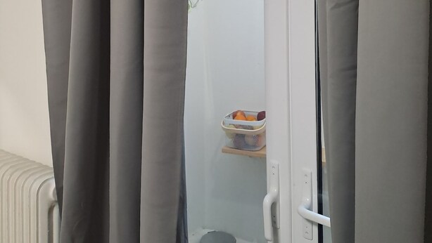 Ein Zimmer mit grauen Vorhängen, einem Heizkörper, einem kleinen Regal mit Obstkorb und einer Wandpflanze.
