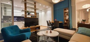 Ein modernes Wohnzimmer mit Flachbildfernseher, blauen Sesseln, einem runden Couchtisch und einem Essbereich mit Stühlen und Tisch.